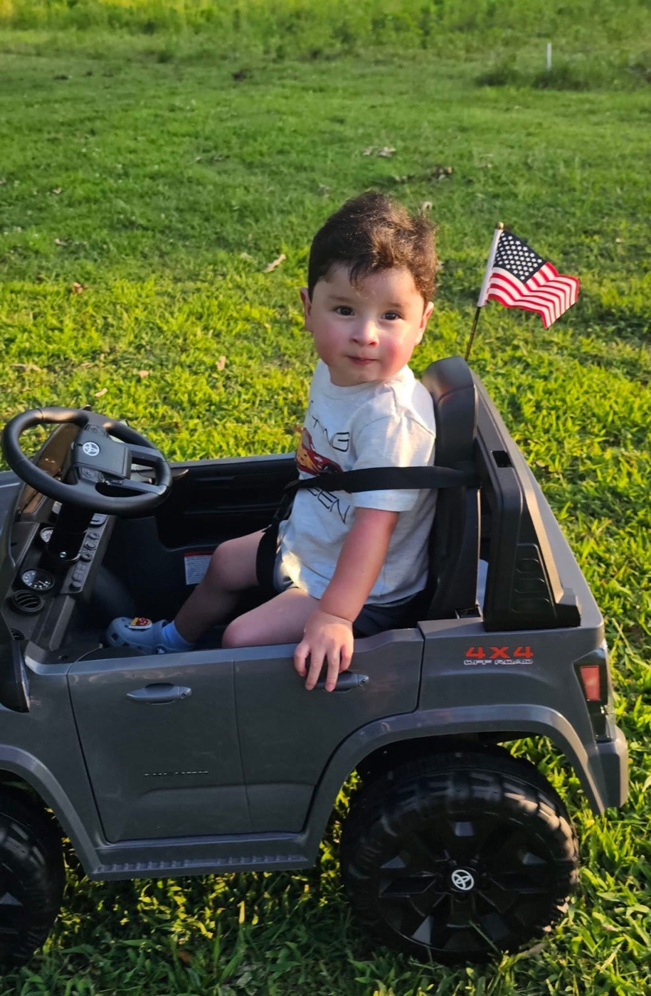 Damian, two years old, sitting behind the wheel of his toy 4x4 in the grass with a small American flag on the back.